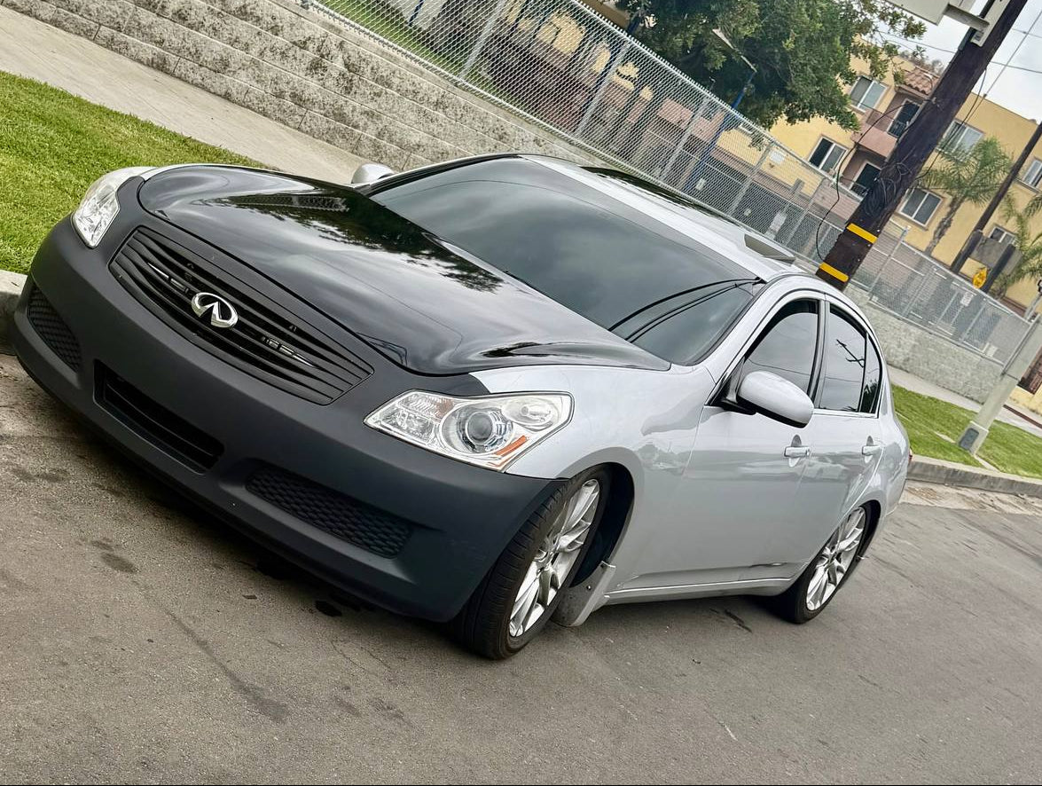 A silver Infiniti G35 sedan parked outdoors on a street with a chain-link fence, trees, and buildings in the background.